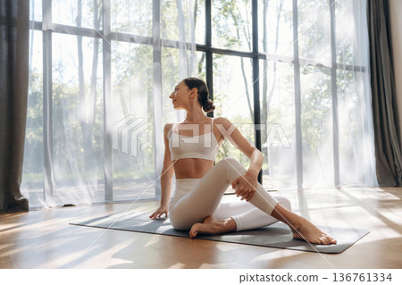 White curtains against her, sitting and relaxing. Young woman in fitness clothes is in the room 136761334