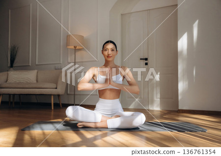 Making yoga gesture by hands, sitting in lotus pose. Young woman in fitness clothes is in the room Making yoga gesture by hands, sitting in lotus pose. Young woman in fitness clothes is in the room 136761354