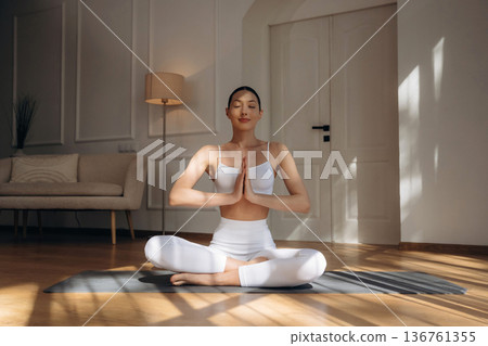 Making yoga gesture by hands, sitting in lotus pose. Young woman in fitness clothes is in the room Making yoga gesture by hands, sitting in lotus pose. Young woman in fitness clothes is in the room 136761355