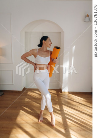 Hobby of yoga, standing and holding the mat. Young woman in fitness clothes is in the room Hobby of yoga, standing and holding the mat. Young woman in fitness clothes is in the room 136761376