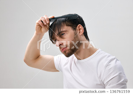 Adult short bearded man in white t shirt combing wet hair forward with black comb, head slightly tilted down, light gray studio background. 136761559