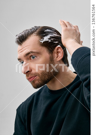 Three quarter view of short bearded man in his thirties in black sweatshirt spreading white foam on side of hair with hand, gray studio background. 136761564