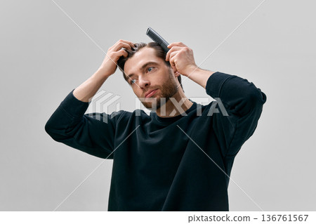 Adult short bearded man in black sweatshirt combing hair with black comb while holding hair with other hand and looking sideway. 136761567