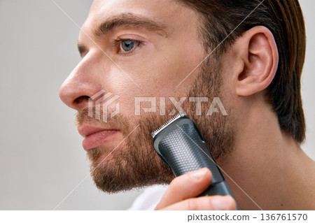 Side close up of adult man trimming short beard with electric trimmer on light gray studio background, facial hair clearly visible Side close up of adult man trimming short beard with electric trimmer on light gray studio background, facial hair clearly visible 136761570