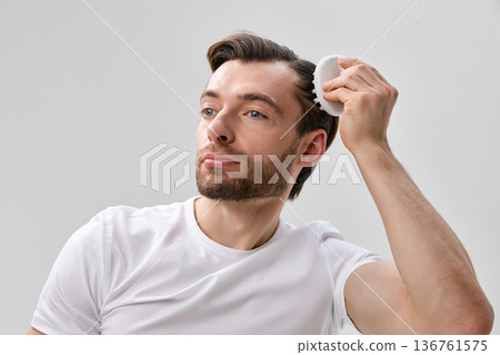 Adult short bearded man in white t shirt using silicone scalp brush on hair while looking forward, on light gray minimal studio background Adult short bearded man in white t shirt using silicone scalp brush on hair while looking forward, on light gray minimal studio background 136761575