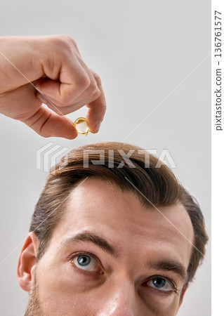 Close up of man applying liquid serum from capsule to hair while looking up on light gray background in minimal studio setting Close up of man applying liquid serum from capsule to hair while looking up on light gray background in minimal studio setting 136761577