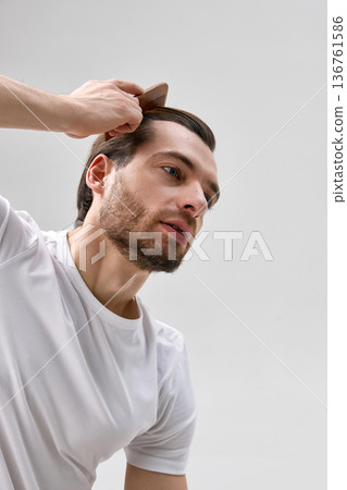Low angle close up of man in his thirties with short beard combing hair with wooden comb on light gray background, 136761586