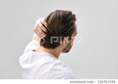 Rear three quarter side view of man in his thirties with short beard touching hair with one hand on light gray background 136761588