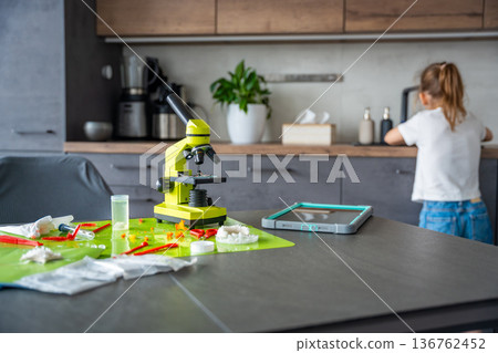 Home science experiment workspace with microscope and tools while child washing hands in background. Responsible STEM learning and laboratory safety at home. Home science experiment workspace with microscope and tools while child washing hands in background. Responsible STEM learning and laboratory safety at home. 136762452