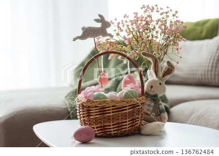 Easter Basket with Ester Eggs, Plush Bunny and Spring Decor on White Table intoCozy Living Room Easter Basket with Ester Eggs, Plush Bunny and Spring Decor on White Table intoCozy Living Room 136762484