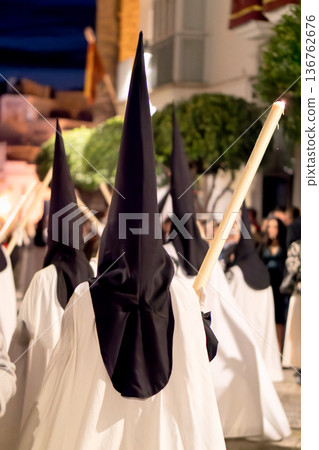 Nazarenes in white tunics and black hoods walking in a night procession with candles. 136762676
