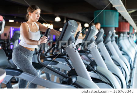 Young woman stands on simulator, performs fitness task using an elliptical simulator Young woman stands on simulator, performs fitness task using an elliptical simulator 136763343
