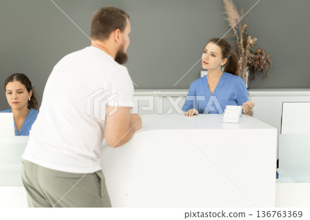 man is standing near the reception in the dentistry and asks about an appointment with a dentist 136763369