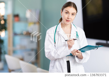 Portrait of young coach female doctor standing with folder with documents and materials in hands and talking in corporate seminar and inviting in auditorium Portrait of young coach female doctor standing with folder with documents and materials in hands and talking in corporate seminar and inviting in auditorium 136763371