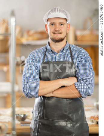 Male baker stands near work table in bakery, resting while working. Male baker stands near work table in bakery, resting while working. 136763460