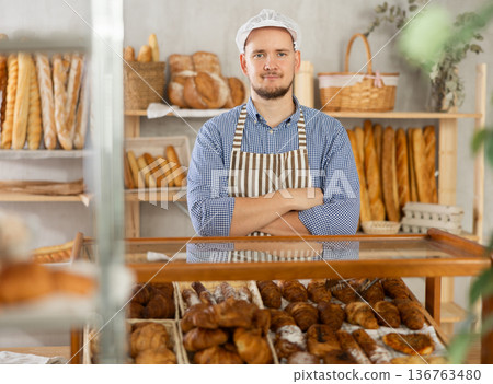 Portrait of a positive baker in an apron against the background of a display case with fresh croissants Portrait of a positive baker in an apron against the background of a display case with fresh croissants 136763480