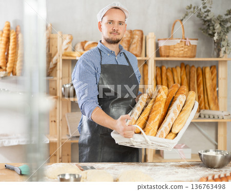 Male baker holds basket with finished products, shows many different baguettes Male baker holds basket with finished products, shows many different baguettes 136763498