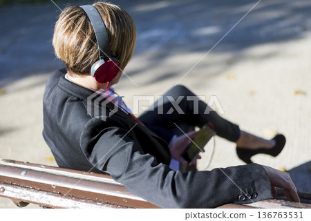 Businesswoman listening to music on park bench Businesswoman listening to music on park bench 136763531