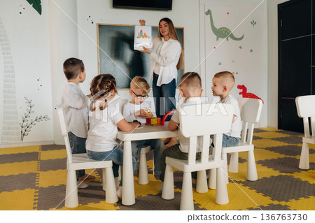 White table, sitting and learning. Children in kindergarten with their teacher White table, sitting and learning. Children in kindergarten with their teacher 136763730