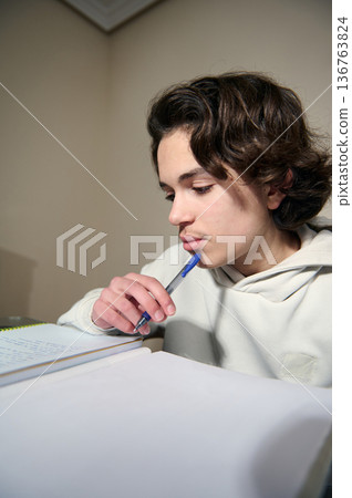 Teenage Boy Studying At Home Doing Homework In Cozy Evening Interior With Pen And Notebooks 136763824