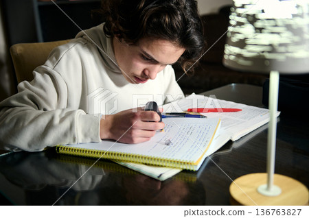 Teenage Boy Doing Homework At Night In Cozy Home Interior With Desk Lamp Teenage Boy Doing Homework At Night In Cozy Home Interior With Desk Lamp 136763827