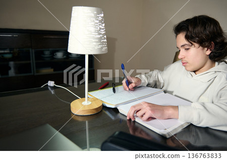 Teenage Boy Studying At Home Doing Homework Under Lamp In Cozy Evening Interior Teenage Boy Studying At Home Doing Homework Under Lamp In Cozy Evening Interior 136763833