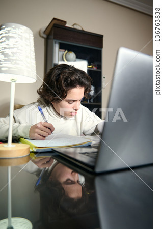 Teenage Boy Doing Homework At A Cozy Home Desk In The Evening With Laptop And Lamp 136763838