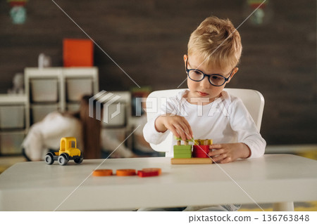 White shirt, eyewear, sitting by table and playing. Group of children are in the kindergarten 136763848