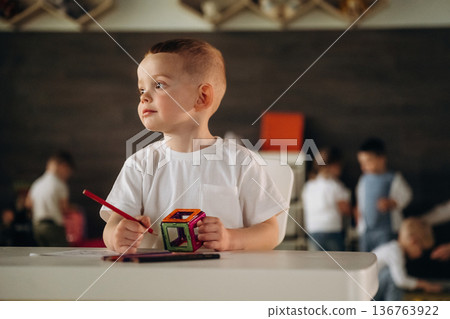 Cute boy is sitting by the table and playing. Group of children are in the kindergarten 136763922