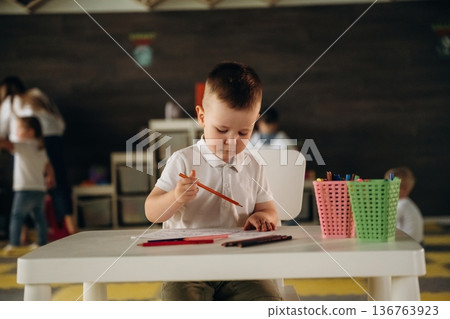 Cute boy is sitting by the table and playing. Group of children are in the kindergarten Cute boy is sitting by the table and playing. Group of children are in the kindergarten 136763923