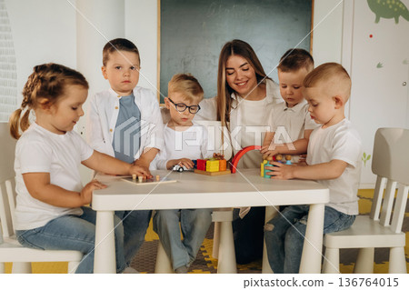 Sitting by the table. Children in kindergarten with their teacher Sitting by the table. Children in kindergarten with their teacher 136764015