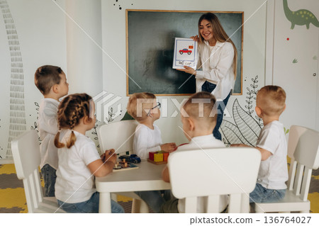 Woman is showing picture of object. Children in kindergarten with their teacher Woman is showing picture of object. Children in kindergarten with their teacher 136764027