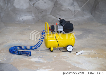 Portable yellow air compressor with a blue coiled hose on a concrete floor in an industrial workshop setting. Portable yellow air compressor with a blue coiled hose on a concrete floor in an industrial workshop setting. 136764315