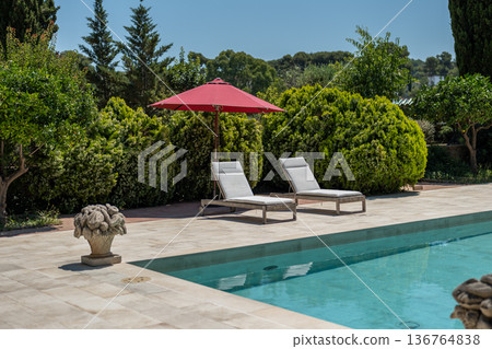 A serene outdoor pool area with two white lounge chairs under a red umbrella. Lush green bushes and trees surround the space, creating a tranquil atmosphere. A serene outdoor pool area with two white lounge chairs under a red umbrella. Lush green bushes and trees surround the space, creating a tranquil atmosphere. 136764838