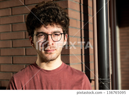 A young man with curly hair and glasses, contemplating a city view from a balcony, with buildings and distant mountains creating an urban background A young man with curly hair and glasses, contemplating a city view from a balcony, with buildings and distant mountains creating an urban background 136765095