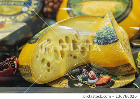 Cheese wheel and cheese wedge wrapped in plastic on store counter with fruit and berries in delicatessen display. Concept of cheese tasting, gourmet food selection, dairy product retail Cheese wheel and cheese wedge wrapped in plastic on store counter with fruit and berries in delicatessen display. Concept of cheese tasting, gourmet food selection, dairy product retail 136766020