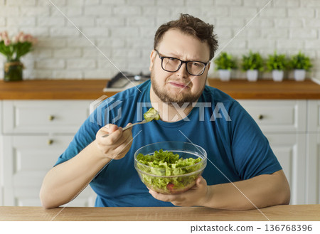 Sad overweight man eating healthy vegetable salad in home kitchen, holding fork 136768396