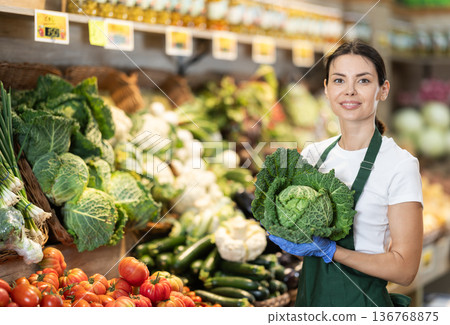 Female grocery store worker offers ripe broccoli cabbage in grocery department of supermarket Female grocery store worker offers ripe broccoli cabbage in grocery department of supermarket 136768875