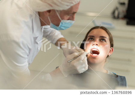 Mature man doctor dentist in white coat examines oral cavity of girl patient in dentistry. 136768896