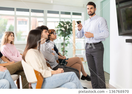 Male lecturer communicates with students, controls projector using remote control. 136768970