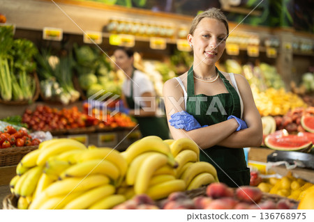 Woman seller in apron and gloves stands in sales area of vegetable store Woman seller in apron and gloves stands in sales area of vegetable store 136768975