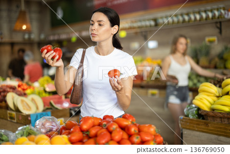 Woman carefully selects fresh tomatoes, standing near the counter in supermarket 136769050