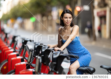 Brunette in blue dress rents electric bike on street of european city 136769101