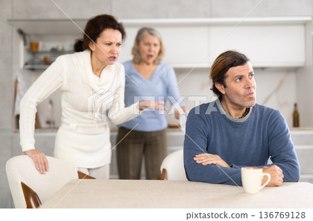 Man turned away from his wife and mother swearing at him in kitchen at home Man turned away from his wife and mother swearing at him in kitchen at home 136769128