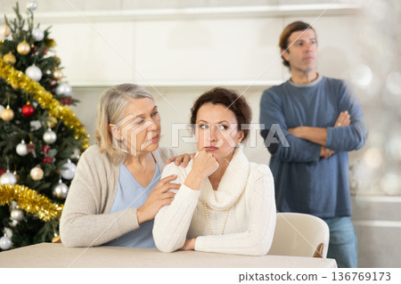 Man on christmas eve quarrels with his wife in kitchen, standing behind her, and elderly mother hugs and calms her daughter Man on christmas eve quarrels with his wife in kitchen, standing behind her, and elderly mother hugs and calms her daughter 136769173