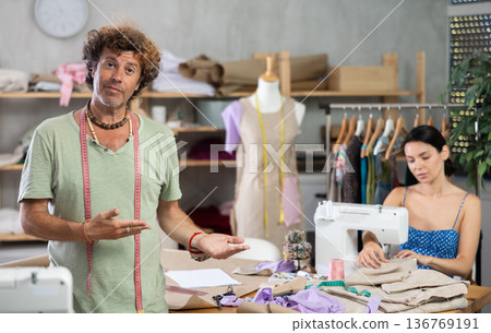 Middle-aged male dressmaker inviting clients while girl sew Middle-aged male dressmaker inviting clients while girl sew 136769191