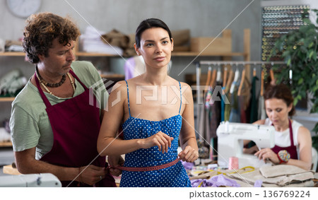 Male dressmaker measuring young woman while assistant sewing Male dressmaker measuring young woman while assistant sewing 136769224