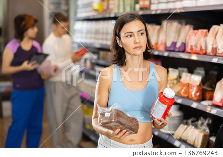 Woman buyer compares glycemic index of bread product and sports nutrition supplement in jar 136769243