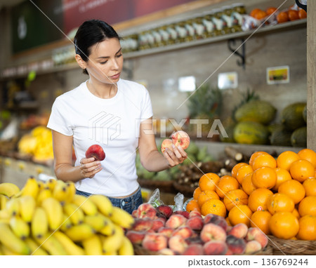 Woman stands in a supermarket near a bountiful fruit counter and chooses a flat peach. Customer prepares to weigh the fruit before buying Woman stands in a supermarket near a bountiful fruit counter and chooses a flat peach. Customer prepares to weigh the fruit before buying 136769244