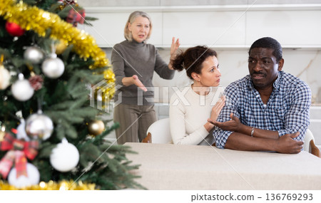 Middle-aged couple sitting at table and mature woman scolding them in the kitchen with Christmas-tree Middle-aged couple sitting at table and mature woman scolding them in the kitchen with Christmas-tree 136769293